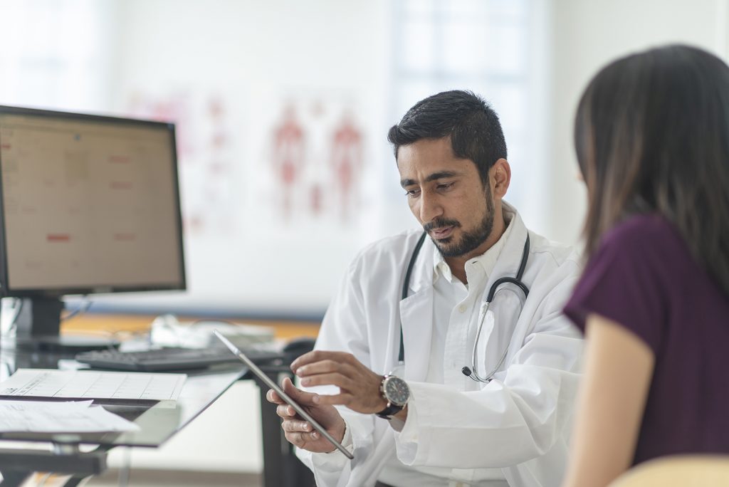 A male doctor of indian descent has a consultation with a patient of hers one afternoon. He is using a tablet to convey the results of her most recent test.